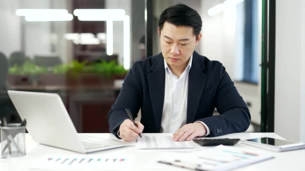 忙碌的亚洲人金融家使用计算器进行财务计算 他坐在工作场所的办公桌前做笔记 有信心的商人穿着正装工作 在商务办公室填写文件 — 图库视频影像