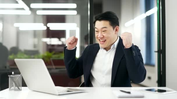 満足している幸せなアジアのビジネスマンは ビジネスオフィスの職場に座っている間 ラップトップで素晴らしいニュースを受け取った後 成功を祝います 興奮笑顔 喜んで成熟した男性 正式なスーツ ダンス 面白いです — ストック動画