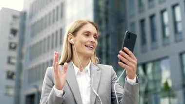 Bir ofis binasının yanında dikilirken telefon ve kulaklıkla konuşan gülümseyen bir iş kadını. Resmi takım elbiseli mutlu bir kadın müşterisiyle ya da iş ortağıyla konuşuyor.