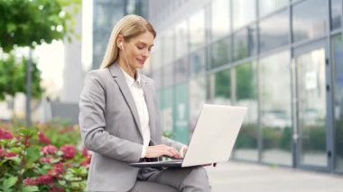 İş kadını bir ofis binasının yanındaki bankta otururken dizüstü bilgisayarda çalışıyor. Resmi takım elbiseli kadın girişimci bir istemciyle çevrimiçi sohbet ediyor, e- postaları kontrol ediyor veya mesaj yazıyor