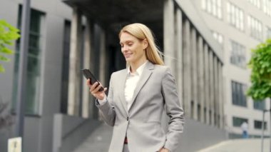 Resmi takım elbiseli mutlu işadamı bir ofis binasının yanında sokakta yürürken telefon kullanıyor. Kadın yönetici bir metin mesajı yazar sohbet, mesajlaşma, e-posta kontrolü veya internette gezinme