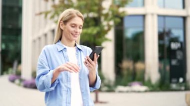 Mutlu sarışın kadın modern bir binanın yanında sokakta yürürken telefonu kullanıyor. Mavi gömlekli olgun bir turist internette mesajlaşarak ya da sosyal medyada gezinerek mesajlaşır.