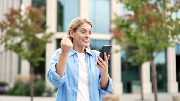 興奮した女性は 近代的な建物の近くの通りに立っている間 彼女が電話で読んだ素晴らしいニュースに満足しています スマートフォンで肯定的な通知を受けた後 成功を祝う笑顔の喜びの女性 — ストック動画