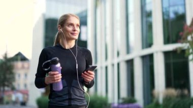 Şehir caddesinde telefonla dolaşan mutlu bir sporcu kadın. Sağlıklı, kulaklıklı, dışarıda müzik dinlemekten hoşlanan bir bayan koşucu. Siyah spor giyimli neşeli bir sporcu antrenmandan dönüyor ya da koşuyor.