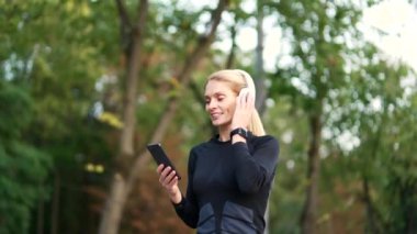 Neşeli, aktif, aktif, kulaklıklı, müzik dinlemekten zevk alan bir kadın. Mutlu gülümseyen kadın şehir parkında koşuyor, akıllı telefondan çalma listesini açıyor ve şarkıdan keyif alıyor.