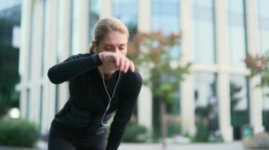 Yetişkin bir bayan koşucu, şehir caddesinde hızlı bir koşudan sonra nefes darlığı çeker. Formda sporcu kadın dizlerinin üzerine çöker alnındaki teri eliyle siler ve derin derin nefes alır. Kapat.