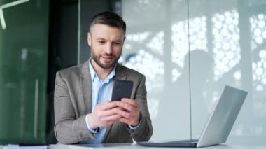 Mutlu iş adamı iş yerindeki iş yerinde akıllı telefon kullanıyor. Sakallı işçi mesajı okur veya yazar, müşteriyle çevrimiçi sohbetler, telefon uygulamalarında bankacılık, e-postaları kontrol etmek veya web tarayıcıları