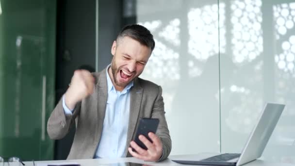令人惊讶的英俊商人坐在商务办公室里 接到了好消息 兴奋快乐的男性员工在智能手机上看到一个愉快的信息 庆祝成功 为胜利而高兴 — 图库视频影像