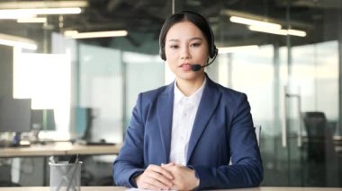 Webcam görüntüsü. Telsiz kulaklıklı Asyalı iş kadını iş yerinde otururken kameraya bakıyor. Kendine güvenen bayan koçun çevrimiçi ya da webinar eğitimi var.