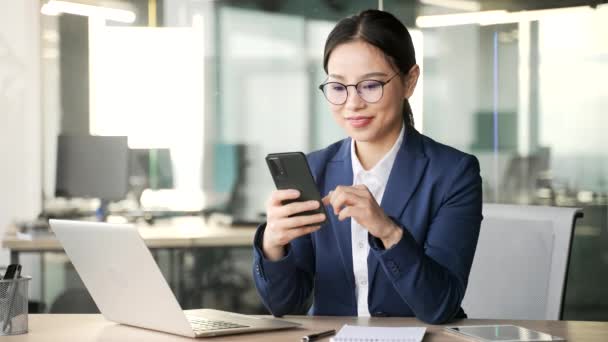 アジアのビジネスマンは ビジネスオフィスで職場に座っている携帯電話を使用しています 正式なスーツの女性従業員は モバイルアプリケーションで動作し メッセージを入力したり クライアントとチャットしたり オンラインで閲覧したりする — ストック動画