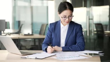 Meşgul Asyalı genç iş kadını iş yerindeki iş yerinde oturan kalemle belgeleri dolduruyor. Kadın girişimci ya da finansör evrak işi yapıyor, mali rapor hazırlıyor ya da vergi formu yazıyor