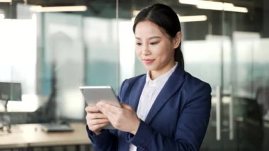 Asyalı iş kadını iş yerinde dikilirken dijital tablet kullanıyor. Resmi takım elbiseli kadın çalışan mobil uygulama üzerinde çalışıyor, yazı okuyor, istemciyle sohbet ediyor veya internette geziniyor