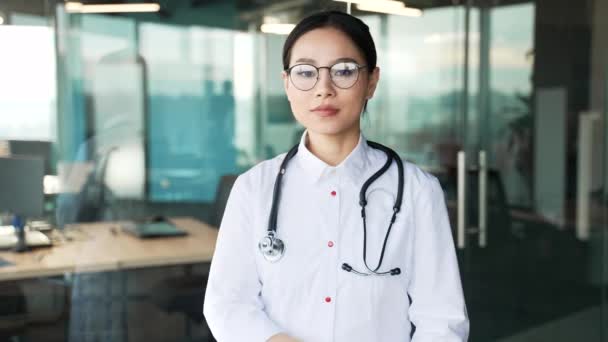 白いコートと現代の病院の診療所でカメラを見ている眼鏡の若い穏やかなアジアの女性医師の肖像画 自信のある女性医療従事者の医師は オフィスに立っている交差した腕でポーズします — ストック動画