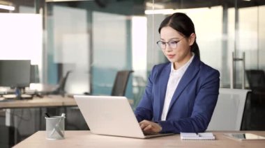 Mutlu Asyalı iş kadını laptopta harika haberler okuyor iş yerindeki iş yerinde oturuyor. Resmi takım elbiseli kadın, başarıyı kutluyor, zafere seviniyor, gülümsüyor, evet işareti yapıyor.