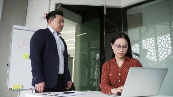成熟したアジアの男性の従業員は ビジネスオフィスの職場で若い女性の同僚を性的に嫌がらせします 大人のビジネスマンは机に座っているショックを受けた女性の同僚の肩に手を置きます — ストック動画