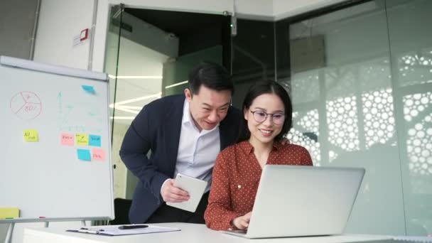 アジアの男性従業員は ビジネスオフィスの職場で若い女性の同僚と浮気します 大人の成熟したビジネスマンは 彼の机に座っている魅力的な女性とロマンス ジョークを開始したいです — ストック動画