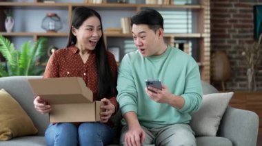Neşeli Asyalı çift müşteri paketini evde açıyor. Mutlu eş ve koca alıcısı kanepeye oturmuş paketleri açıyor. E-ticaretin tatmin olmuş müşterisi. İyi geri bildirim. Ekspres dağıtım hizmetleri 