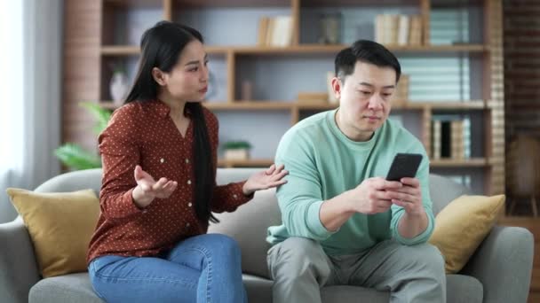 愤怒的亚洲妻子和丈夫坐在客厅的沙发上争吵 一个女人在和一个漠不关心的男人争吵 这个男人看手机 沉迷于社交媒体和互联网 — 图库视频影像
