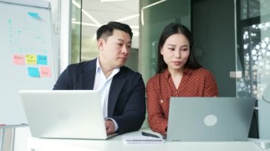 İki Asyalı iş arkadaşı iş konularını tartışıyor, iş yerindeki işyerinde danışmanlık yapıyorlar. Erkek yönetici, genç kadın uzmana yardım eder. Takım çalışması. Çalışanlar konuşur, fikirleri dizüstü bilgisayarlarla açıklar.