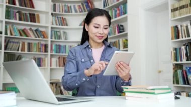 Asyalı kız öğrenci kampüs kütüphanesinde otururken dijital tablet kullanarak öğreniyor. Genç bir kadın internette sohbet ediyor ya da üniversitenin sınav dönemine hazırlanırken internette geziniyor.