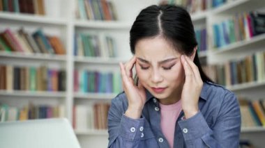 Baş ağrısı çeken Asyalı bir kız öğrenci kampüs kütüphanesinde dizüstü bilgisayarla çalışıyor. Genç kadın masajını bozar, alnına ve şakaklarına masaj yapar. Fazla mesai çalışmaktan çok yoruldu. Kapat.