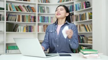 Alerjisi olan Asyalı bir kız öğrenci kampüs kütüphanesinde otururken dizüstü bilgisayarda hapşırıyor. Hasta genç bir kadın burnunu mendille siler. Soğuk algınlığı, virüs ya da grip belirtileri gösteriyor.