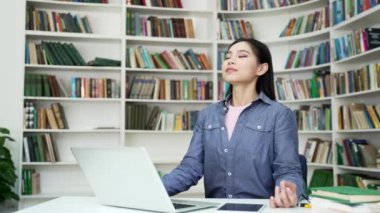 Genç Asyalı kız öğrenci, kampüs kütüphanesinde otururken gözleri kapalı meditasyon yapıyor. Genç kadın laptopta çalışmak için çalışmaya ara verdi. Sakin kız dinleniyor, rahatlıyor, huzuru hissediyor.
