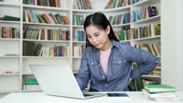坐在校园图书馆的笔记本电脑上工作时 亚洲的女学生背痛得很厉害 这让她很不安 年轻女人按摩后背 按摩脊椎肌肉 她有坐骨神经痛或冷神经 — 图库视频影像