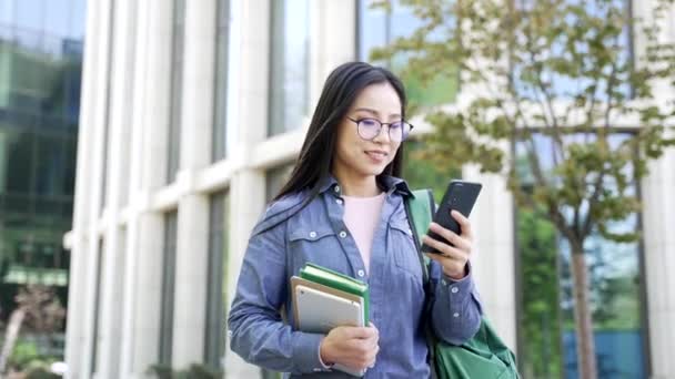 大学ビルの近くのキャンパススペースを歩く携帯電話を使用して笑顔のアジアの女性学生 バックパックと手の本を持つ女性は メッセージを読み ソーシャルネットワークを閲覧し 友人とオンラインチャットします — ストック動画