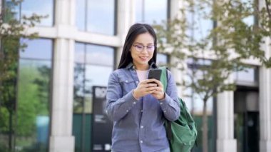 Mutlu Asyalı kız öğrenci, üniversite binasının yakınındaki sokakta telefonla harika haberler okuyarak başarıyı kutluyor. Gülümseyen genç kadın sevinçli bir mesaj aldı, olumlu bir uyarı.