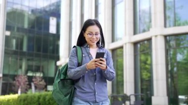 Gülümseyen Asyalı kız öğrenci üniversite binasının yanındaki kampüste telefon kullanarak yürüyor. Sırt çantalı mutlu bir kız mesajları okur, sosyal ağlara göz atar ya da arkadaşıyla internetten sohbet eder.