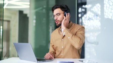 Kablosuz kulaklıklı mutlu iş adamı iş yerinde otururken müzik dinliyor. Neşeli sakallı adam cep telefonundaki çalma listesini açıyor ve iş yerinde şarkı söylemekten hoşlanıyor.