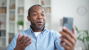 Olgun Afro-Amerikalı bir adam video konuşmasında selfie çekiyor ve evdeki oturma odasındaki kanepede oturan akıllı telefon ekranına bakıyor. Gülümseyen yaşlı sakallı bir erkek arkadaşıyla internette sohbet ediyor.