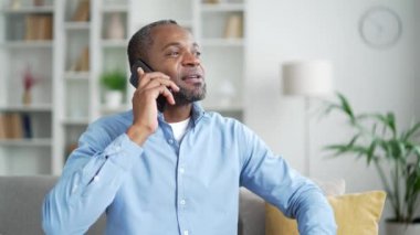 Evdeki oturma odasında kanepede otururken akıllı telefondan konuşan mutlu Afro-Amerikan erkeği. Gülümseyen yaşlı sakallı bir erkek bir meslektaşıyla konuşur ya da bir arkadaşıyla iletişim kurar.