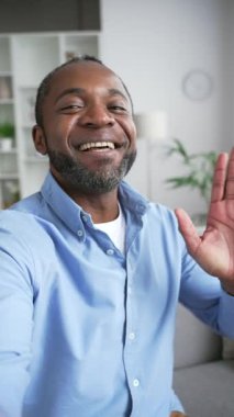 POV dikey video. Olgun Afro-Amerikalı bir adam video konuşmasında akıllı bir telefonla konuşuyor. Evdeki kanepede oturan kameraya bakıyor. Gülümseyen kıdemli işadamı iş ortağıyla internette sohbet ediyor.