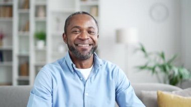 Olgun Afro-Amerikalı adam parmaklarıyla imza atıp evdeki kanepede oturan kameraya bakıyor. Gülümseyen kıdemli siyahi adam tamam sembolünü göster, her şeyi iyi gösterir, başarı jesti, onaylama