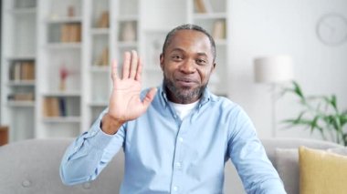 Webcam görüntüsü. Gülümseyen Afro-Amerikan iş adamı kameraya bakarak konuşuyor. Oturma odasındaki kanepede oturuyor. Mutlu bir serbest çalışanın müşterisiyle uzak bir görüşmesi olur.