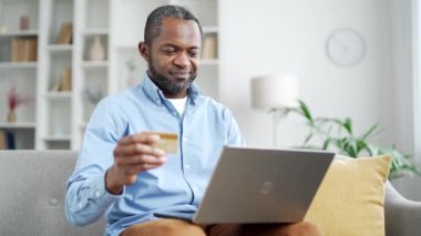 Mutlu Afro-Amerikalı adam online alışveriş yapıyor dizüstü bilgisayarında kredi kartı numarasını yazıyor evdeki kanepede oturuyor. Gülümseyen yaşlı sakallı erkek satın alıyor, terfi ve indirimlerle tatmin oluyor.