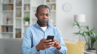 Kafası karışmış bir Afro-Amerikan erkeği evdeki koltukta otururken akıllı telefon kullanmakta zorlanıyor. Endişeli erkeğin telefonda çalışırken ve kameraya bakarken karmaşık bir sorunu var.