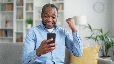 Mutlu Afro-Amerikalı adam akıllı telefondan harika haberler aldı evdeki oturma odasındaki kanepede oturuyor. Gülümseyen yaşlı sakallı erkek telefonda hoş bir mesaj okuyor, başarıyı kutluyor, evet, vay canına.
