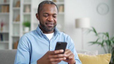 Olgun bir Afro-Amerikan erkeği evdeki oturma odasındaki koltukta akıllı telefon kullanıyor. Gülümseyen erkek sosyal ağlara takılır, çevrimiçi alışveriş yapar, bir arkadaşıyla sohbet eder, mesaj yazar veya okur. Kapat.