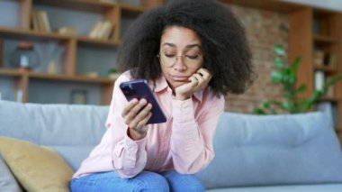 Yalnız, üzgün, genç Afrikalı Amerikalı kadın evdeki oturma odasında kanepede otururken akıllı telefon kullanmaktan sıkıldı. Uykulu siyah kadın depresyonda ve kötü bir ruh halinde, cep telefonuyla sosyal medyada geziniyor.