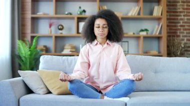 Sakin genç Afro-Amerikan kadın Lotus pozisyonunda meditasyon yaparken gözleri kapalı evdeki oturma odasında kanepede oturuyor. Mutlu siyah kadın yoga yapıyor, derin derin nefes alıyor, rahat bir koltukta dinleniyor.