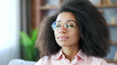 Genç, gülümseyen Afro-Amerikan kadın gözleri kapalı bir şekilde evdeki kanepede oturuyor. Sakin, mutlu, gözlüklü, dinlenen, derin derin nefes alan ve kameraya bakan siyah kadın.