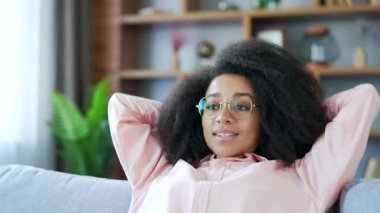Genç, gülümseyen Afro-Amerikan kadın. Evdeki oturma odasında kanepede otururken gözleri kapalı ve elleri başının arkasında dinleniyor. Sakin, mutlu, gözlüklü siyah kadın işten sonra dinleniyor.
