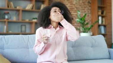 Genç Afrikalı Amerikalı kadın evdeki oturma odasında kanepede otururken ağrı kesici alıyor ve bardaktan su içiyor. Aşırı yorgun siyah kadın baş ağrısı çekiyor ve kafasına masaj yapıyor.