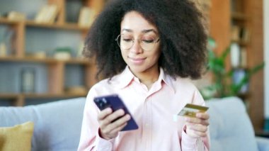 Mutlu Afro-Amerikan kadın online alışveriş yapıyor. Telefonla kredi kartı numarasını yazıyor. Evdeki kanepede oturuyor. Gülümseyen siyah kadın satın alıyor. İndirimlerden memnun. Kapat.