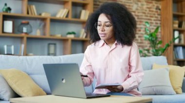 Genç Afro-Amerikalı kız öğrenci evde oturan bir dizüstü bilgisayarla online e-öğrenme, video kursu dinliyor. Uzaktan öğrenim seminerinde ya da eğitimde olan siyahi bir kadın öğretmenle defter konuşmaları yazıyor.