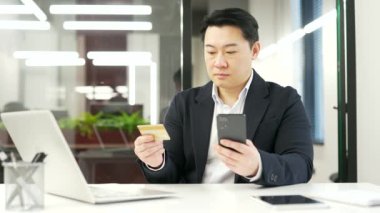 Asyalı işadamı, bir iş yerinde kredi kartı ve telefon kullanarak online alışveriş yapıyor. Resmi takım elbiseli bir adam siparişler ve alımlar yapar, indirimler ve terfiler konusunda mutludur.