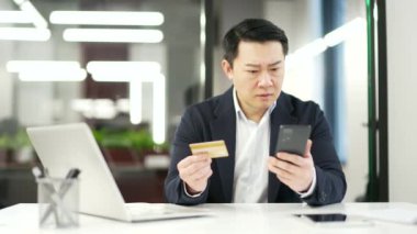 Şok olmuş Asyalı adam iş yerinde otururken kredi kartını telefonla kontrol ediyor. Hayal kırıklığına uğramış erkek dolandırıcılıkla karşı karşıya, hesabından para çalınmış. O bir aldatma kurbanı oldu.